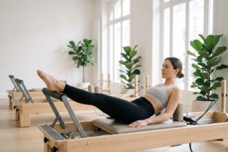 Femme pratiquant le Pilates reformer dans un studio parisien moderne, allongée sur le chariot avec les jambes tendues sur la barre, tenue de sport noire ajustée